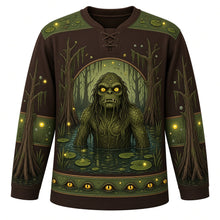 Load image into Gallery viewer, Ugly Halloween Swamp Creature Bayou Hockey Jersey in Bog Brown, front view – bog brown body with a scaly swamp creature with glowing eyes rising from a murky bayou crest with moss-draped cypress trees, lily pads, fireflies and bubbles, moss yoke, lily pad sleeve bands and a cypress tree hem band – custom name and number