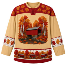 Load image into Gallery viewer, Ugly Thanksgiving Covered Bridge Foliage Hockey Jersey in Birch Gold, front view – birch gold body with barn red yoke, a red wooden covered bridge with a gray roof over a rocky creek among white birch trunks and round fluffy autumn trees, oak leaves on the water, rounded oak-leaf yoke, birch-bark sleeve bands with oak leaves and a hem band of little red covered bridges among fall trees – custom name and number
