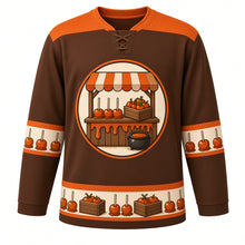 Load image into Gallery viewer, Ugly Thanksgiving Caramel Apple Stand Hockey Jersey in Barnwood Brown, front view – barnwood brown body with pumpkin orange yoke with a round crest of a rustic wooden farm stand with a striped awning and a counter of glossy caramel apples on sticks, a crate of apples and a caramel pot, caramel-drip yoke, caramel-apple sleeve bands and a caramel-apple-and-crate hem band – custom name and number